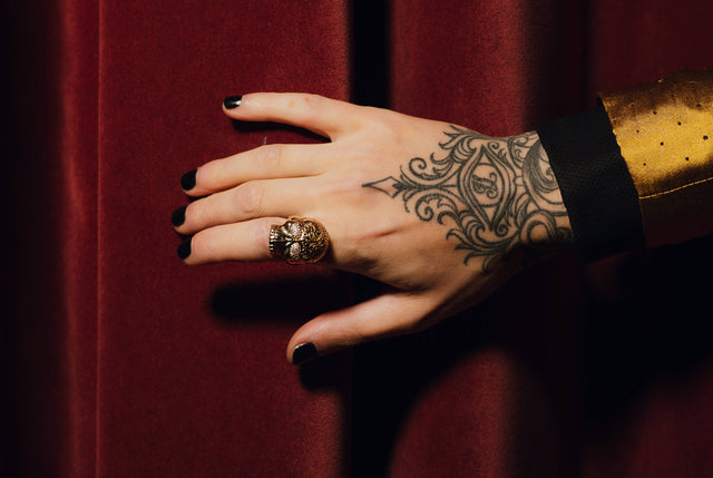 Man's hand with a tattoo in front of a red curtain is wearing a gold skull ring with diamond pave eyes and a tattoo pattern on the skull's head.